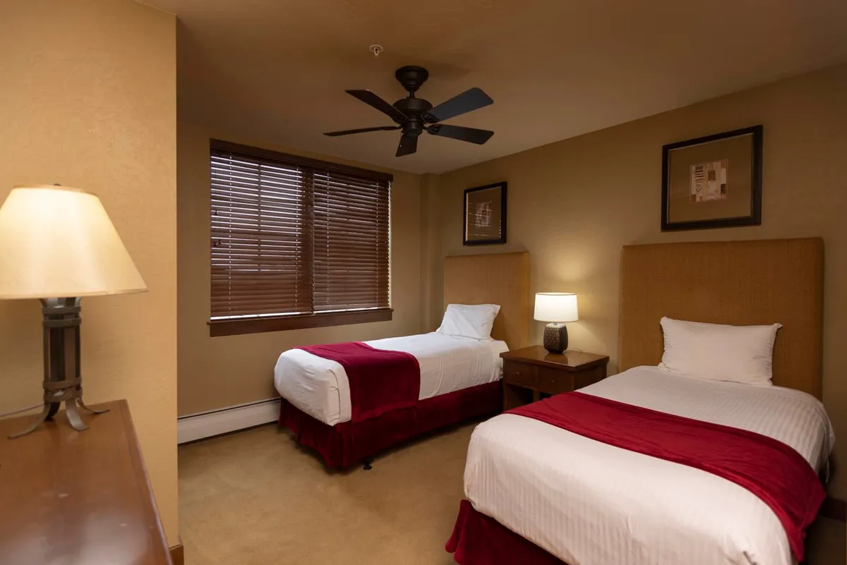 The scene shows a bedroom with two twin beds, each with red bed skirts, a window allowing natural light, and a table lamp. The space is practical for families or friends, offering comfort and flexibility.
