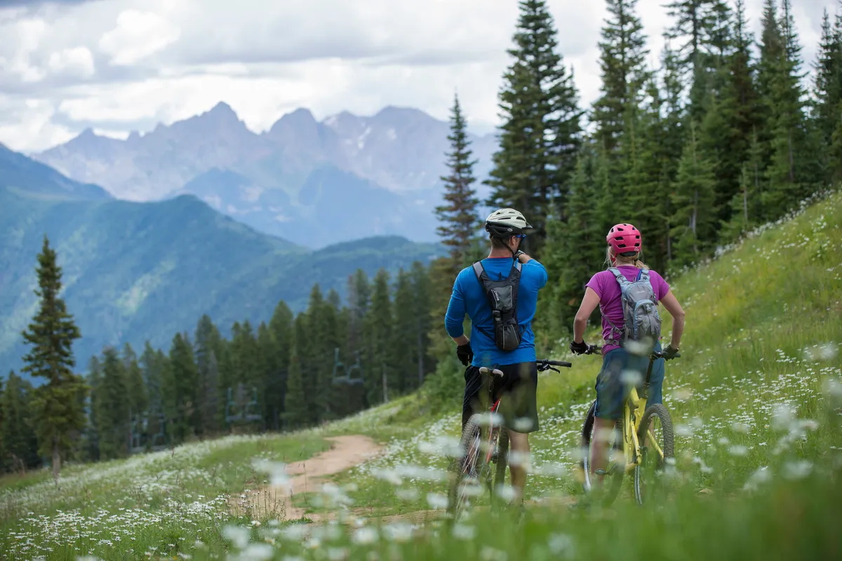 An expansive mountain vista forms the backdrop for two mountain bikers riding along a wildflower-lined path beneath a vibrant treeline. The image exudes a sense of adventure and connection to nature, capturing the thrill of outdoor activities available in Purgatory Village.