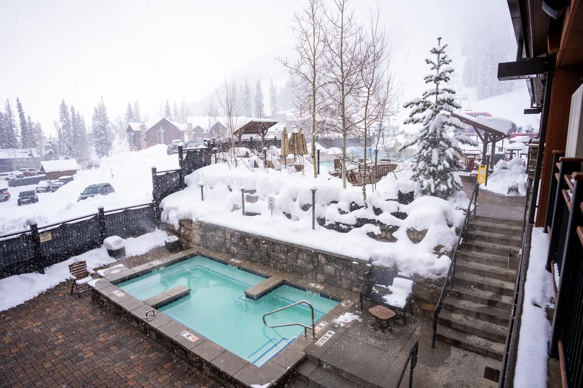 A serene outdoor hot tub sits against a snowy backdrop, surrounded by trees and a sturdy staircase, inviting guests to soak and unwind while snow falls gently around them. The image conveys warmth and tranquility, ideal for après-ski relaxation.