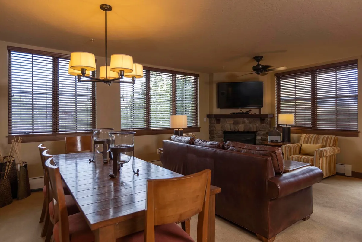 A cozy living space featuring a leather sofa, stone fireplace, flat-screen TV, and large windows with window treatments. The dining table is set for family meals, and the space feels inviting and comfortable for gatherings or relaxing evenings.