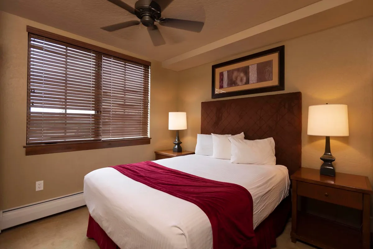 A tranquil bedroom with a plush bed, soft bedside lamps, and a window with blinds partially open to let in mountain light. Decorative artwork creates a homey atmosphere ideal for unwinding after a day outdoors.