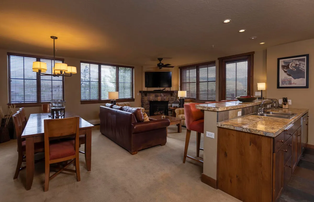 A wide view of an open-concept living and dining area features a stone-faced gas fireplace, comfortable living room seating, a generous dining table, and a kitchen island. Large windows reveal glimpses of the mountains outside, and the arrangement is ideal for relaxing, entertaining, or gathering with friends and family.