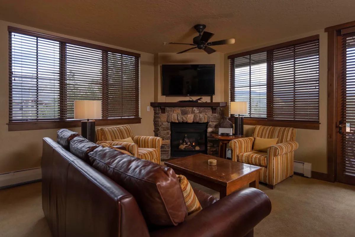 A welcoming living room featuring a comfortable leather couch, stone gas fireplace, wooden coffee table, and striped armchairs. Large windows allow ample natural light and offer views of the surrounding landscape, setting the scene for relaxation and warmth.