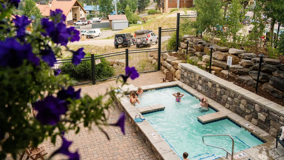 This image captures a lively outdoor swimming pool surrounded by blooming flowers, with several people enjoying the water and relaxing by the poolside. The inviting atmosphere is enhanced by the presence of families and friends, suggesting a communal and playful environment perfect for unwinding after adventurous days.