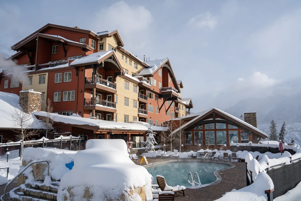 An exterior winter scene showing the lodge's large building, an outdoor pool, and snow-covered surroundings. The building’s balconies face the mountains, suggesting outdoor spaces to enjoy crisp air and sunsets even in winter.