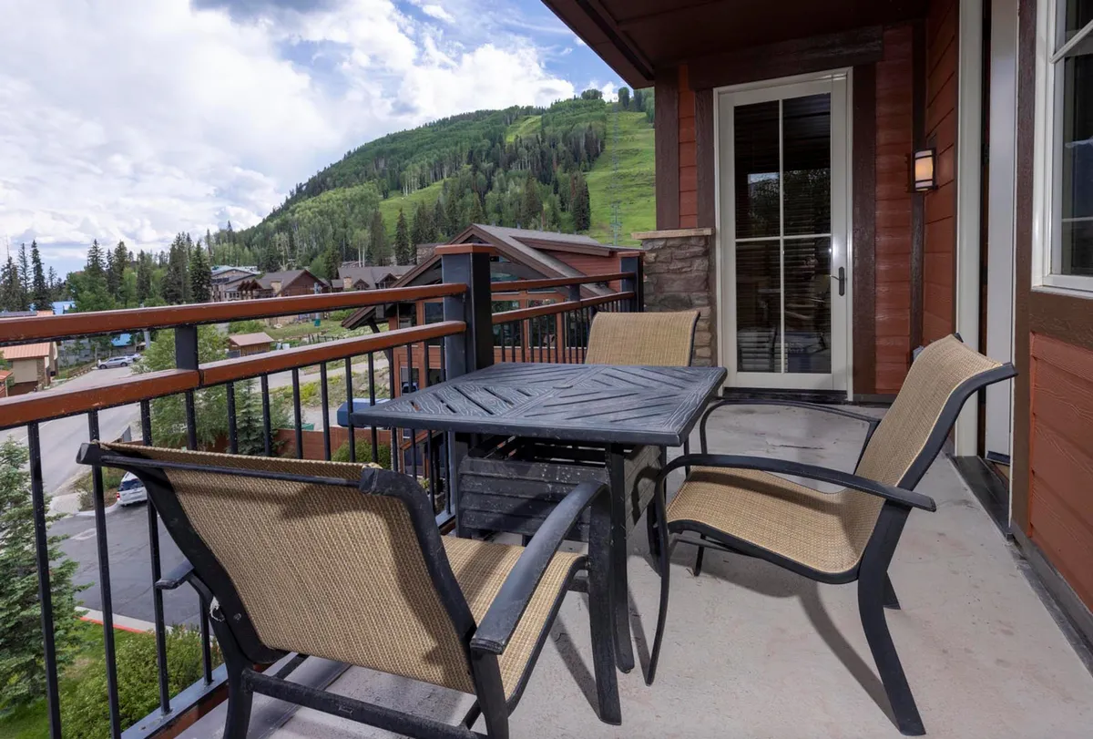 The image features a private patio with a table and chairs overlooking breathtaking mountain vistas, surrounded by lush trees. The view showcases a tranquil mountain landscape, possibly at sunset or dawn, with hints of morning mist and vibrant foliage that change with the seasons. The scene invites relaxation and connection with nature, ideal for activities like sipping coffee, unwinding after a day on the slopes, or stargazing under the clear Colorado sky.