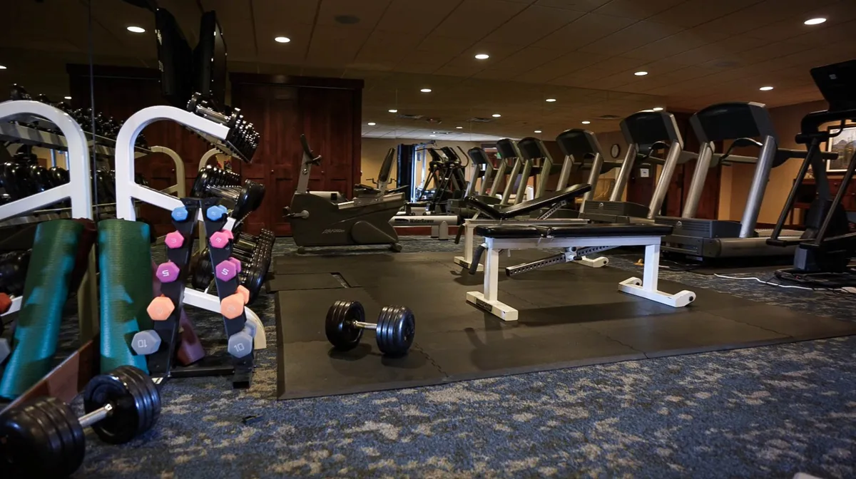 The image shows a bright, well-equipped fitness center with dumbbells, treadmills, a weight bench, and exercise mats. The space is inviting, with ample room for personal workouts or group fitness activities. The lighting and motivational atmosphere suggest a place to energize both body and mind.