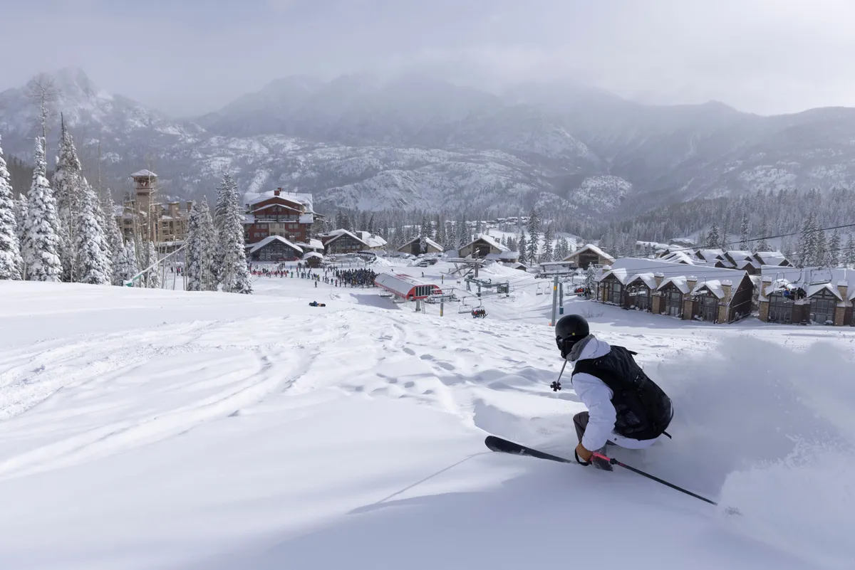 A dynamic shot of a skier gliding down a snowy slope, framed by snow-laden trees and mountains in the background. The ski resort buildings are visible, highlighting the close relationship between the accommodations and the slopes.